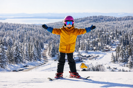 Ett barn står på skidor med armarna utsträckta i en snöklädd skidbacke, med snötäckta skogar och bebyggelse i bakgrunden.