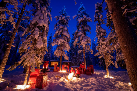 ”Snöklädd vinterkväll i skogen med människor klädda som jultomtar. De sitter vid en brasa och är omgivna av stora inslagna paket och upplysta granar.”
